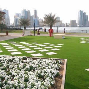 Dubai landscape design featuring lush green lawns, decorative flower beds, paved walkways, palm trees, and a waterfront city skyline.