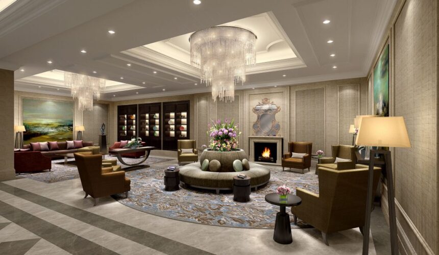 Elegant hotel lobby lounge with crystal chandeliers, a circular tufted sofa, leather armchairs, fireplace with ornate mirror, and floral centerpiece on a patterned round rug.