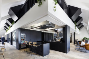 Modern office breakout area with a black kitchenette featuring a beverage fridge, espresso machine, and wooden bar stools, set beneath angular black staircases with hanging plants, herringbone wood flooring, and a lounge seating area in the background.