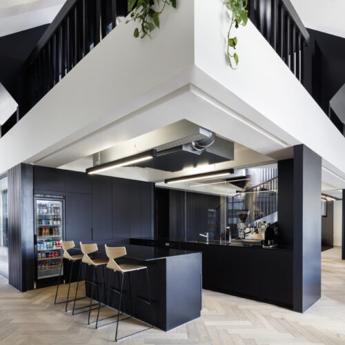 Modern office breakout area with a black kitchenette featuring a beverage fridge, espresso machine, and wooden bar stools, set beneath angular black staircases with hanging plants, herringbone wood flooring, and a lounge seating area in the background.
