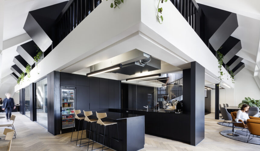 Modern office breakout area with a black kitchenette featuring a beverage fridge, espresso machine, and wooden bar stools, set beneath angular black staircases with hanging plants, herringbone wood flooring, and a lounge seating area in the background.