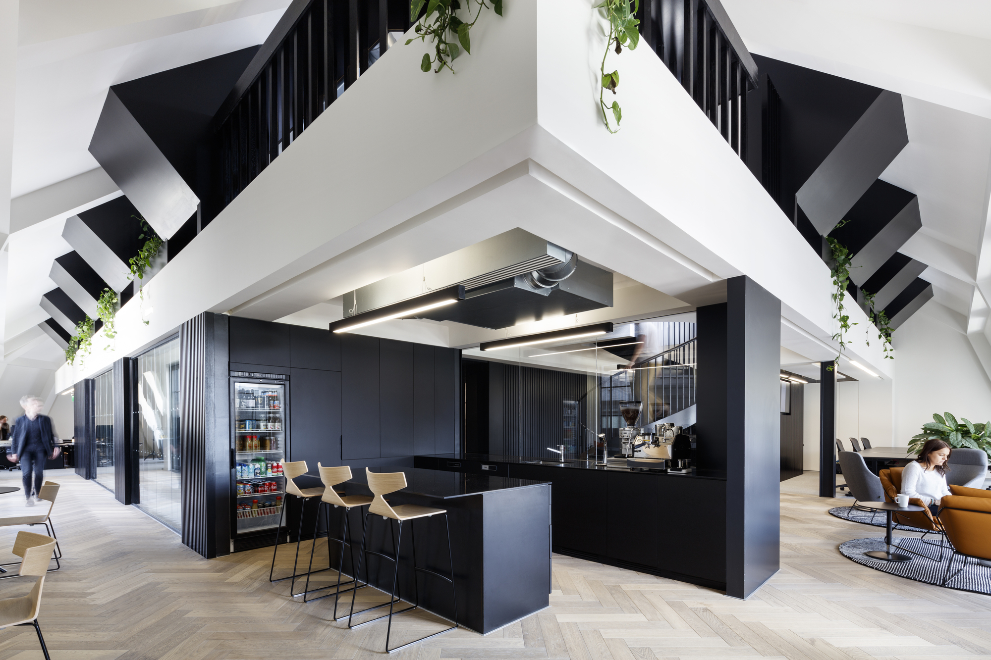 Modern office breakout area with a black kitchenette featuring a beverage fridge, espresso machine, and wooden bar stools, set beneath angular black staircases with hanging plants, herringbone wood flooring, and a lounge seating area in the background.