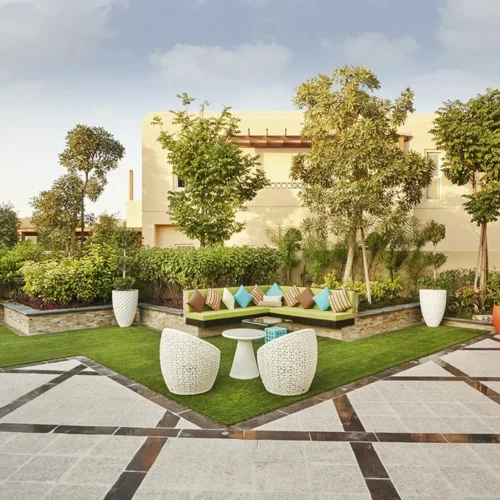 Landscaped villa courtyard with a green corner sofa and colorful cushions, white woven chairs and table on artificial turf, surrounded by trees, orange planters, and a patterned stone-tiled patio.