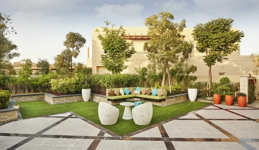 Landscaped villa courtyard with a green corner sofa and colorful cushions, white woven chairs and table on artificial turf, surrounded by trees, orange planters, and a patterned stone-tiled patio.