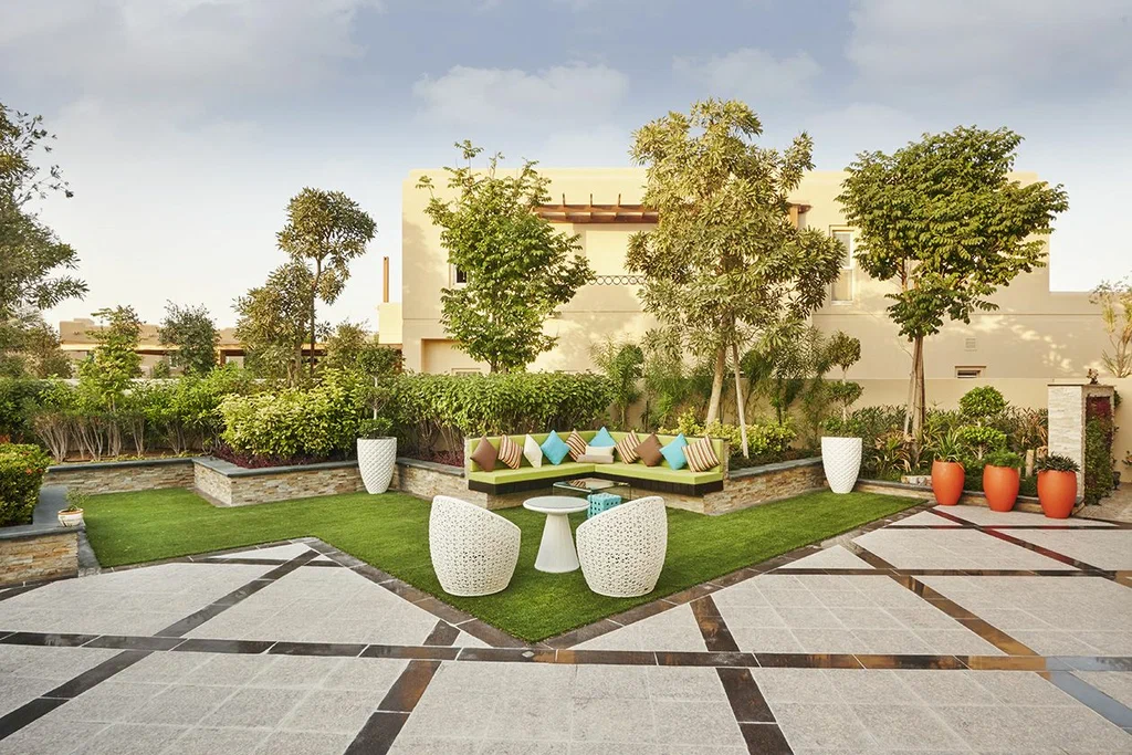 Landscaped villa courtyard with a green corner sofa and colorful cushions, white woven chairs and table on artificial turf, surrounded by trees, orange planters, and a patterned stone-tiled patio.