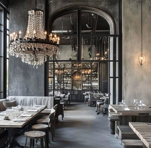 Industrial-chic restaurant interior with grey concrete walls, a black crystal chandelier, wooden tables and stools, upholstered banquette seating, arched mirrored bar area, and large steel-framed windows.